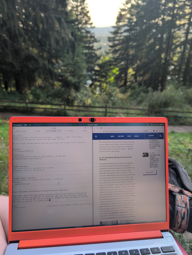 An orange Framework laptop held open in a sunlit pine forest, screen split between a Pi coding agent terminal on the left and a web browser showing a wweek.com article about Portland&rsquo;s best cheap eats on the right.