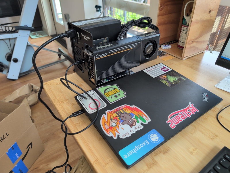 A sticker-covered ThinkPad laptop on a wooden desk connected via Thunderbolt to a black external GPU dock. The Dock has an AMD Radeon AI Pro R9700 GPU and a PC power supply.